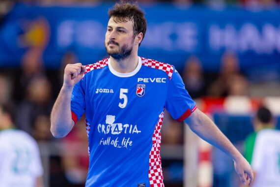 Ein männlicher Handballspieler im blauen Kroatien-Trikot mit rot-weiß karierten Schultern hebt während eines Spiels jubelnd die Faust, im Hintergrund sind unscharfe Spieler und eine Menschenmenge zu sehen.