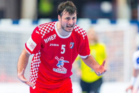 Ein männlicher Handballspieler in einem rot-weiß karierten Trikot mit der Nummer 5 gestikuliert während eines Spiels mit offenen Händen und intensivem Blick. Im Hintergrund sind andere Spieler und ein Tor zu sehen.