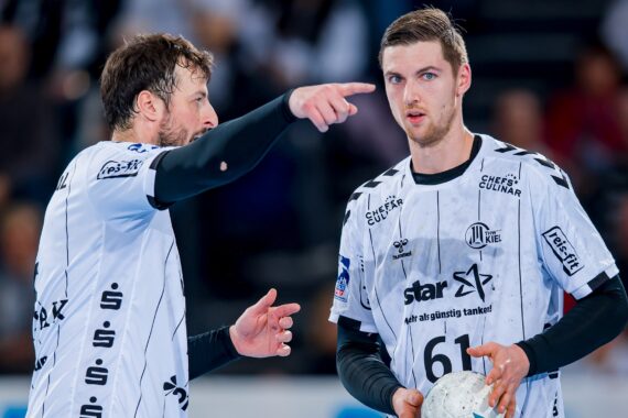 Zwei männliche Handballspieler in weißen Trikots mit schwarzen Details befinden sich auf dem Spielfeld. Ein Spieler zeigt auf etwas und gibt Anweisungen, während der andere den Ball hält und aufmerksam zuhört. Beide scheinen während des Spiels konzentriert zu sein.