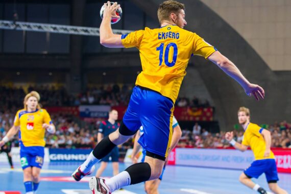 Ein männlicher Handballspieler in einem gelben Trikot springt in die Luft, um den Ball zu werfen. Im Hintergrund sind andere Spieler und Zuschauer bei einem Hallenspiel zu sehen.