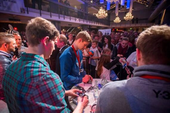 Eine Gruppe junger Männer gibt Autogramme an einem Tisch, während sich eine Menge Fans, darunter auch Kinder, in einer hell erleuchteten Halle mit Kronleuchtern versammeln.
