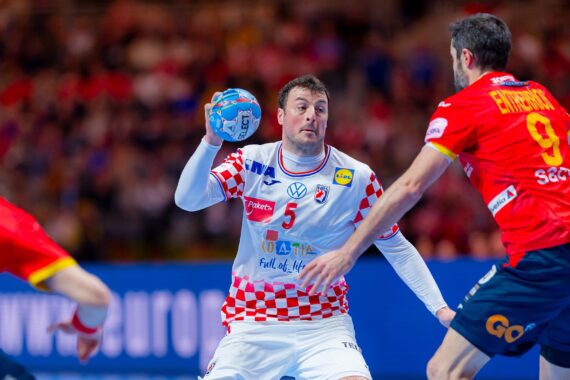 Ein männlicher Handballspieler in einem weiß-rot karierten Trikot bereitet sich darauf vor, den Ball zu werfen, während zwei gegnerische Spieler in rot-gelben Trikots ihn während eines Spiels verteidigen.
