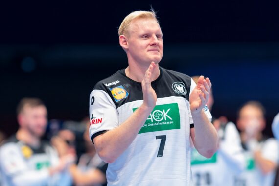 Ein männlicher Handballspieler, der ein weißes Trikot mit der Nummer 7 trägt, klatscht in die Hände und steht auf einem Spielfeld, während die anderen Mannschaftsmitglieder im Hintergrund verschwommen zu sehen sind.