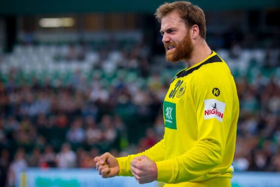 Ein männlicher Handballspieler in einem gelben Trikot ballt die Fäuste und jubelt auf einem Hallenplatz, während im Hintergrund unscharfe Zuschauer zu sehen sind.