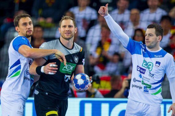 Drei männliche Handballspieler in Aktion; einer in Schwarz hält den Ball, eng bewacht von zwei Spielern in weiß-blauen Trikots, während der Spieler rechts den Daumen nach oben streckt. Im Hintergrund sind Zuschauer zu sehen.