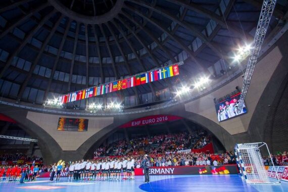 Weitwinkelansicht einer Handballhalle während der EHF Euro 2016, mit Mannschaften auf dem Spielfeld, Zuschauern auf den Tribünen und Flaggen verschiedener Länder über dem Spielfeld.