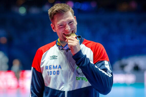 Ein männlicher Sportler in einer rot-weiß-blauen Jacke hält eine Goldmedaille in die Höhe und lächelt, während er in einer Sporthalle mit unscharfem blauem Hintergrund steht.