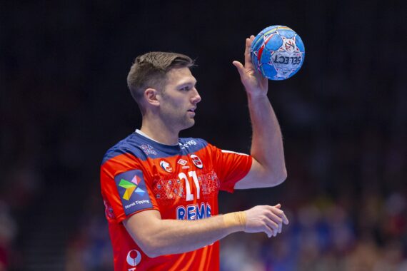 Ein männlicher Handballspieler in einem orange-blauen Trikot hält einen blauen Handball in einer Hand und bereitet sich auf den Wurf vor. Der Hintergrund ist unscharf, um die Konzentration auf den Sportler hervorzuheben.