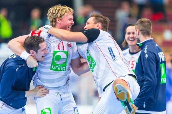 Vier männliche Handballspieler in weiß-blauen Trikots feiern fröhlich auf dem Spielfeld, umarmen sich und lächeln, während die Fans im Hintergrund verschwimmen. Ihre Trikots sind mit Sponsorenlogos versehen.