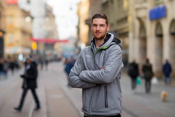 Ein Mann mit hellbraunem Haar und Bart steht selbstbewusst mit verschränkten Armen auf einer Stadtstraße und trägt eine graue Jacke. Unscharfe Fußgänger und historische Gebäude säumen den Hintergrund.