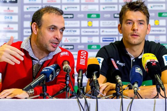 Zwei Männer sitzen an einem mit Mikrofonen bedeckten Pressekonferenztisch. Der eine im roten Pullover spricht und gestikuliert, der andere im schwarzen Hemd hört zu, im Hintergrund sind die Logos der Sponsoren zu sehen.
