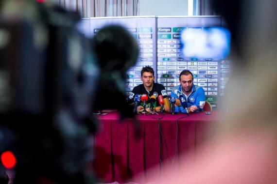 Zwei Männer sitzen während einer Pressekonferenz an einem Tisch mit Mikrofonen und sind Reportern und Kameras zugewandt. Im Hintergrund sind Sponsorenlogos zu sehen, und der Vordergrund ist durch ein Kameraobjektiv unscharf.