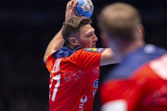 Ein männlicher Handballspieler in einem roten Trikot bereitet sich darauf vor, den Ball zu werfen, während ein anderer Spieler im Vordergrund steht. Der Fokus liegt auf seinem entschlossenen Gesichtsausdruck und seinem erhobenen Arm, der den blau-weißen Ball hält.