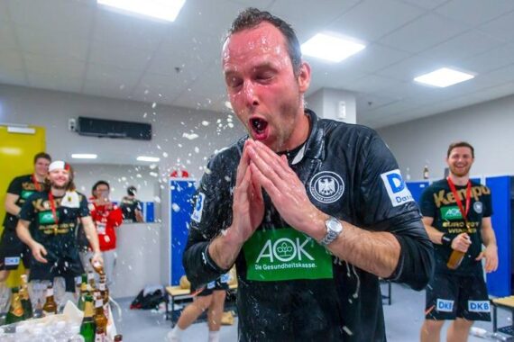 Ein Mann in einer Sportuniform steht in einer Umkleidekabine, durchnässt und mit gefalteten Händen, während er zur Feier des Tages mit Flüssigkeit übergossen wird. Andere Mannschaftskameraden im Hintergrund lächeln und halten Flaschen in der Hand.