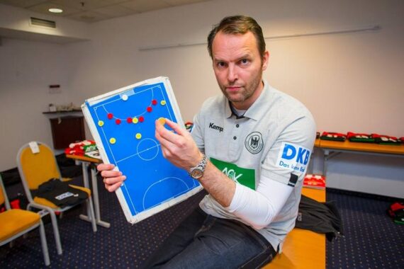 Ein Mann in einer Sportuniform sitzt in einem Raum und hält eine magnetische Taktiktafel mit farbigen Spielsteinen in der Hand, auf der er eine Strategie erklärt. Im Hintergrund sind auf Stühlen und Tischen Sporttrikots ausgelegt.