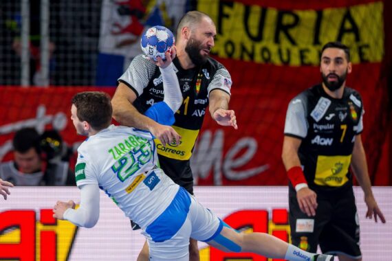 Ein Handballspieler in einem schwarzen Trikot bereitet sich darauf vor, den Ball zu werfen, während er während eines Spiels von einem Gegner in einem weiß-blauen Trikot eng verteidigt wird. Im Hintergrund sind Zuschauer und Fahnen zu sehen.