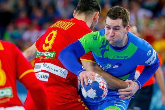Ein männlicher Handballspieler in einem blau-grünen Trikot stürmt mit dem Ball nach vorne, während er während eines Spiels von zwei Gegnern in roten Trikots eng verteidigt wird. Das Spiel ist intensiv und temporeich.