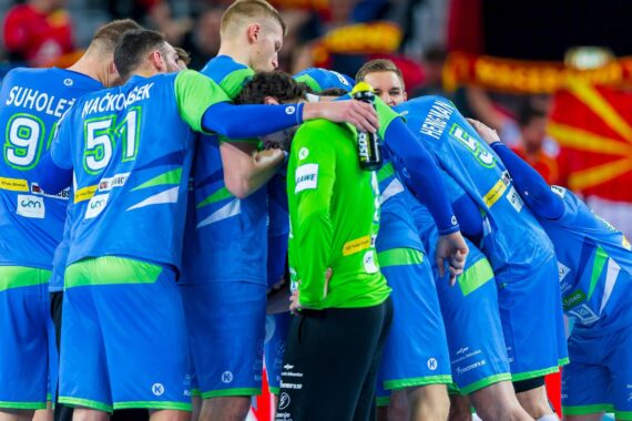 Eine Gruppe von männlichen Handballspielern in blauen und grünen Trikots drängt sich auf einem Hallenplatz eng zusammen und zeigt Teamwork und Konzentration vor einem Spiel. Im Hintergrund ist eine Menschenmenge mit roten und gelben Fahnen zu sehen.