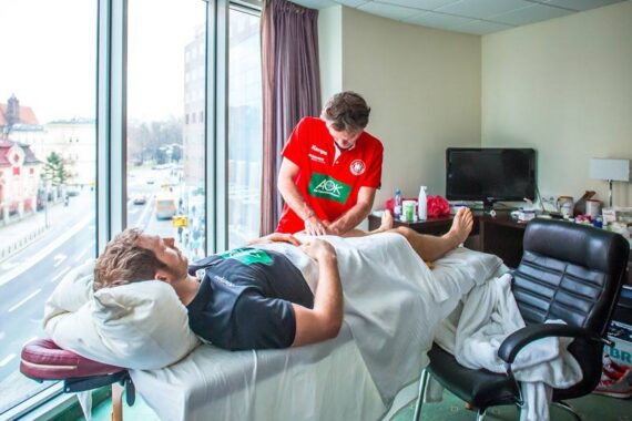 Ein Mann in einem roten Hemd gibt einem anderen Mann, der auf einer Massageliege in einem Raum mit großen Fenstern mit Blick auf eine Stadtstraße liegt, eine Massage oder Physiotherapie.