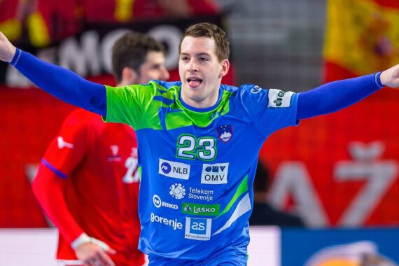 Ein Handballspieler in einem blau-grünen Trikot mit der Nummer 23 jubelt mit erhobenen Armen und herausgestreckter Zunge. Ein anderer Spieler in einem roten Trikot ist im Hintergrund zu sehen.