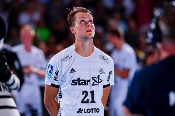 Ein männlicher Handballspieler in einem weißen Trikot mit der Nummer 21 steht auf einem Spielfeld und blickt nach oben. Er ist umgeben von unscharfen Mitspielern, Offiziellen und Kameras, was auf eine professionelle Sportveranstaltung hindeutet.
