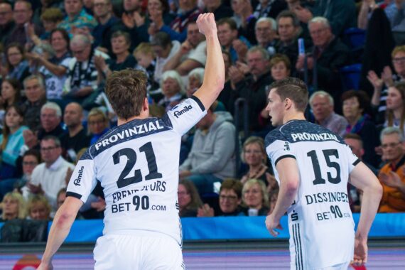 Zwei männliche Handballspieler in weißen Trikots, mit den Nummern 21 und 15, stehen vor einer jubelnden Menge. Spieler 21 hebt jubelnd die Faust, während Spieler 15 zuschaut. Das Publikum sitzt in einer Sportarena.