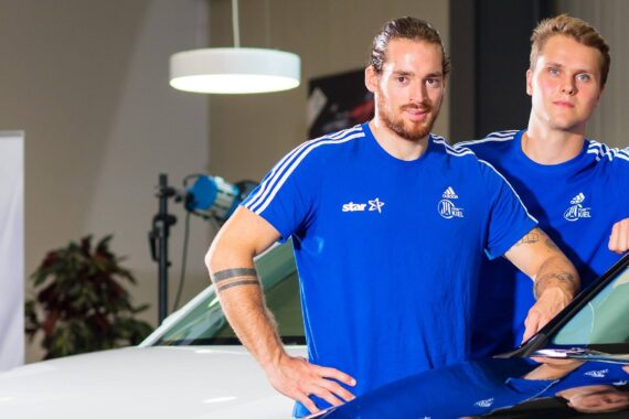 Zwei junge Männer in blauen Sporthemden stehen nebeneinander und lehnen sich an ein Auto in einer Halle. Der eine hat lange Haare und einen Bart, der andere hat kurzes blondes Haar. Beide schauen in die Kamera, im Hintergrund stehen eine Hängelampe und Pflanzen.