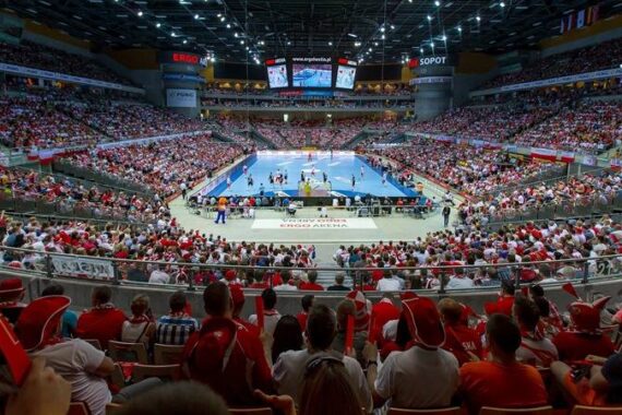 Eine große Sporthalle, gefüllt mit Zuschauern, die ein Handballspiel verfolgen. Die Zuschauer, von denen viele rot und weiß gekleidet sind, umringen das hell erleuchtete Spielfeld. Banner und Fahnen schmücken die Halle.