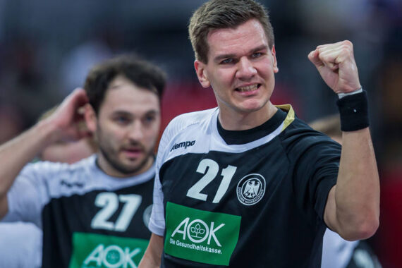 Ein männlicher Handballspieler in einem schwarz-weißen Trikot mit der Nummer 21 jubelt mit geballter Faust. Ein anderer Mitspieler, der die Nummer 27 trägt, steht leicht unscharf hinter ihm.