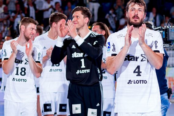 Vier männliche Handballspieler in weißen Trikots und einer in schwarzer Kleidung applaudieren auf einem Spielfeld, während im Hintergrund eine Menschenmenge und Kameras zu sehen sind. Die Spieler sehen fröhlich und feierlich aus.