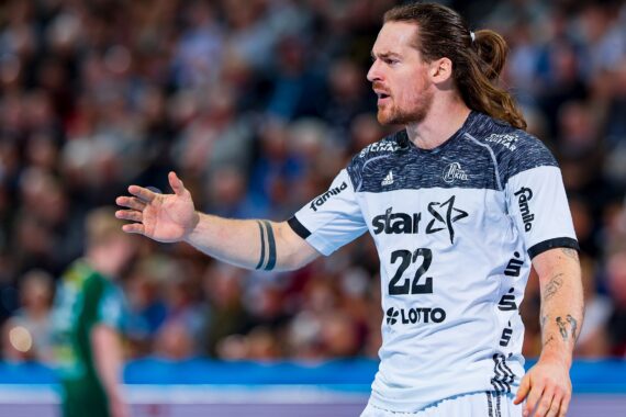 Ein männlicher Handballspieler mit langen Haaren in einem Pferdeschwanz, der ein weiß-schwarzes Trikot mit der Nummer 22 trägt, gestikuliert mit seiner linken Hand während eines Spiels. Im Hintergrund sind ein weiterer Spieler und eine unscharfe Menschenmenge zu sehen.