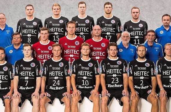 Eine Handballmannschaft der Männer stellt sich für ein Gruppenfoto auf, die Spieler tragen schwarze Trikots, die Betreuer blaue Hemden. Die Mannschaft steht und kniet in Reihen, die Logos sind auf ihren Trikots und an der Wand hinter ihnen zu sehen.