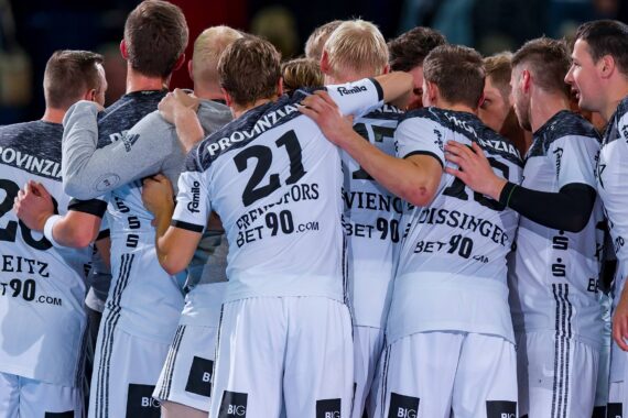 Eine Gruppe männlicher Sportler in weißen und schwarzen Trikots versammelt sich vor einem Spiel auf einem Hallenplatz, um Teamwork und Einigkeit zu zeigen. Auf ihren Trikots stehen Sponsoren, Namen und Nummern.