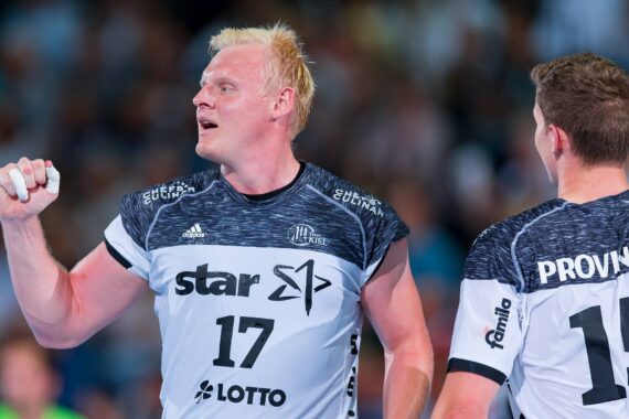 Zwei männliche Handballspieler in schwarzen und weißen Trikots stehen auf einem Spielfeld. Der Spieler mit der Nummer 17, der nach vorne schaut, hält einen Handball und wirkt konzentriert. Der andere Spieler, Nummer 13, ist von hinten zu sehen. Der Hintergrund ist mit Zuschauern unscharf.