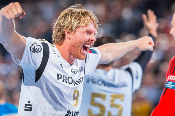 Ein blonder männlicher Handballspieler in einem weißen Trikot mit der Nummer 9 jubelt mit erhobenen Armen und vor Begeisterung geöffnetem Mund, während ein anderer Spieler in einem roten Trikot ihm gegenübersteht. Andere Mannschaftskameraden sind im unscharfen Hintergrund zu sehen.