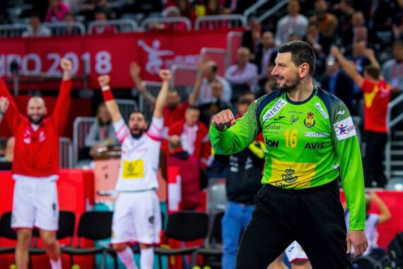 Ein männlicher Handballtorhüter in einem grünen Trikot lächelt und ballt die Faust zum Jubel, während Teamkollegen und Fans im Hintergrund mit erhobenen Armen jubeln. Die Szene ist energiegeladen, und ein rotes Banner der EURO 2018 ist zu sehen.