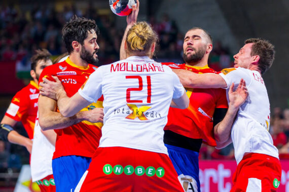 Vier männliche Handballspieler in Aktion, zwei in roten und zwei in weißen Trikots, kämpfen intensiv um den Ball. Ein Spieler in Rot greift nach oben, während die anderen blockieren oder versuchen, den Ball während eines Spiels zu greifen.