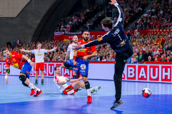 Ein Handballspieler in Blau und Rot schießt auf das Tor zu, während der Torwart in Schwarz springt, um den Ball abzufangen. Andere Spieler und Zuschauer beobachten das intensive Spiel in einer hell erleuchteten Halle.