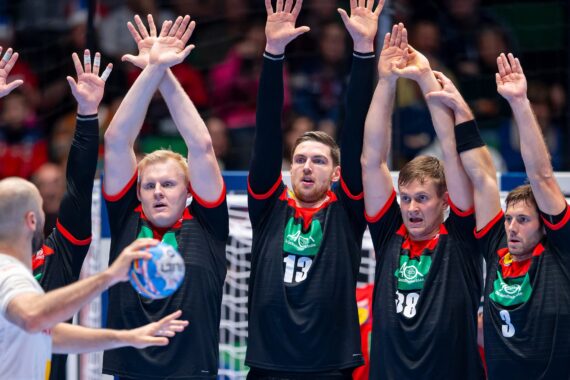 Vier männliche Handballspieler in schwarzen Trikots stehen mit erhobenen Armen nebeneinander und bereiten sich darauf vor, einen Schuss abzublocken, während ein gegnerischer Spieler den Ball hält und zum Wurf bereit ist. Hinter ihnen ist ein Tor zu sehen.
