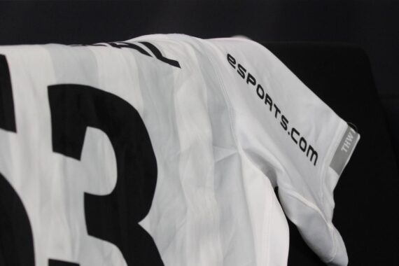Ein weißes Sporttrikot mit der Nummer 3 in schwarzem Fettdruck und dem Schriftzug eSPORTS.COM auf dem Ärmel ist über eine schwarze Fläche drapiert.