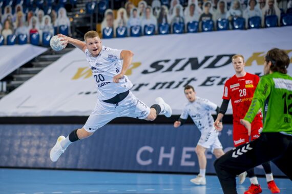 Ein Handballspieler in einem weißen Trikot springt und bereitet sich darauf vor, den Ball in Richtung Tor zu werfen, während der gegnerische Torwart in Grün und Spieler in roten Trikots zusehen und sich auf die Verteidigung vorbereiten. Zuschauer oder Ausschnitte füllen die Plätze im Hintergrund.