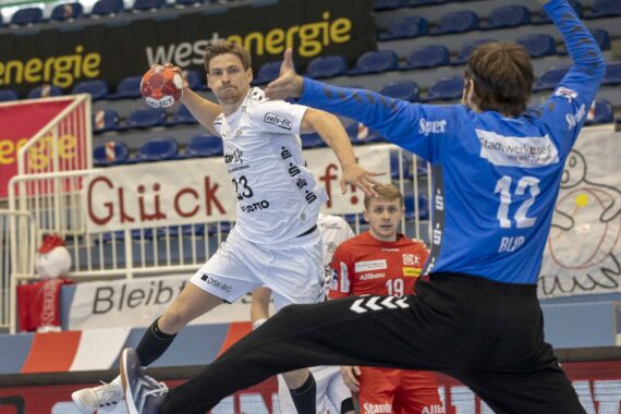 Ein Handballspieler in einem weißen Trikot springt, um den Ball in Richtung Tor zu schießen, während ein Torwart in Blau versucht, den Schuss zu blocken. Ein weiterer Spieler in Rot und leere Plätze sind im Hintergrund zu sehen.