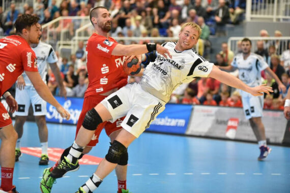 Ein Handballspieler in einem weißen Trikot versucht zu schießen, während er von zwei Spielern in roten Trikots während eines intensiven Spiels auf einem Hallenplatz verteidigt und festgehalten wird. Die Zuschauer schauen von den Tribünen im Hintergrund zu.