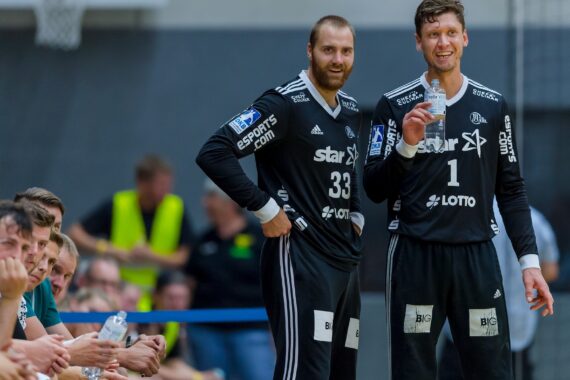 Zwei lächelnde männliche Handballspieler in schwarzen Trikots stehen zusammen auf dem Spielfeld, während ihre Mannschaftskameraden am Spielfeldrand sitzen und zusehen. Die Spieler wirken entspannt und blicken in Richtung Publikum.