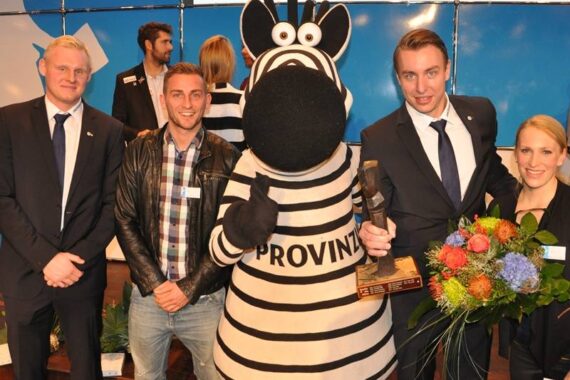 Vier lächelnde Erwachsene stehen neben einer Person in einem schwarz-weiß gestreiften Zebra-Maskottchenkostüm. Einer hält einen Blumenstrauß und ein anderer eine Trophäe in der Hand. Sie scheinen bei einer Indoor-Veranstaltung zu feiern.