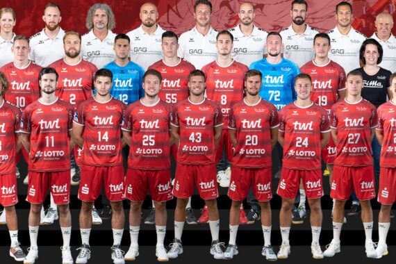 Eine Herren-Handballmannschaft stellt sich für ein Gruppenfoto auf. Die Spieler in roten Trikots stehen vorne, während Trainer und Betreuer in weißen Hemden hinter ihnen stehen, alle mit dem Gesicht nach vorne vor einem roten Hintergrund.