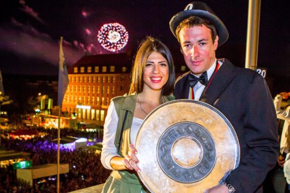 Eine lächelnde Frau und ein Mann in Anzug und Hut halten eine große silberne Trophäenplatte vor einer nächtlichen Menschenmenge, während ein Feuerwerk den Himmel erhellt und die Gebäude im Hintergrund beleuchtet sind.