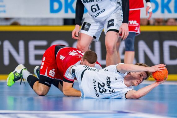 Ein Handballspieler in einem weißen Trikot springt auf das Spielfeld und greift nach einem orangefarbenen Ball, während ein Spieler in einem roten Trikot hinter ihm auf dem Boden steht. Andere Spieler, die teilweise sichtbar sind, stehen in der Nähe.