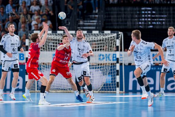 Eine Gruppe männlicher Handballspieler in rot-weißen Trikots kämpft auf einem Hallenplatz um den Ball, während im Hintergrund ein Tor und Zuschauer zu sehen sind.