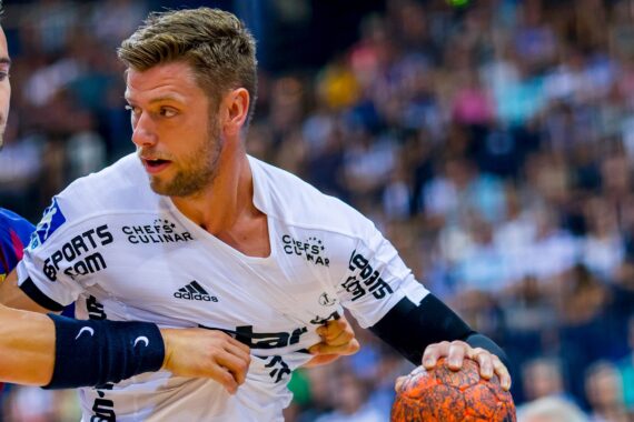 Zwei männliche Handballspieler in Aktion; einer in einem weißen Trikot hält einen orangefarbenen Ball, während er von einem Gegner in einem dunklen Trikot eng verteidigt wird. Der Hintergrund zeigt eine unscharfe Menschenmenge in einem Stadion.
